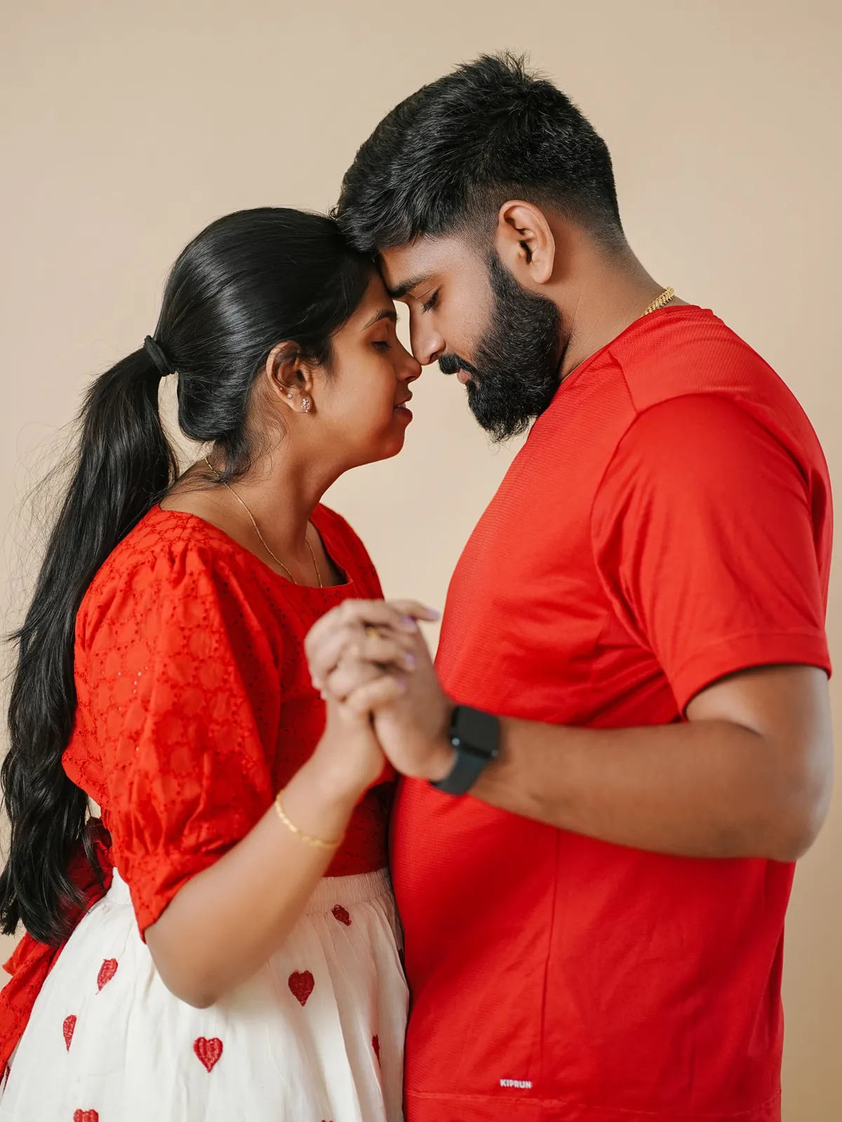 A couple in red stand forehead to forehead with eyes closed and hands clasped, against a beige backdrop.