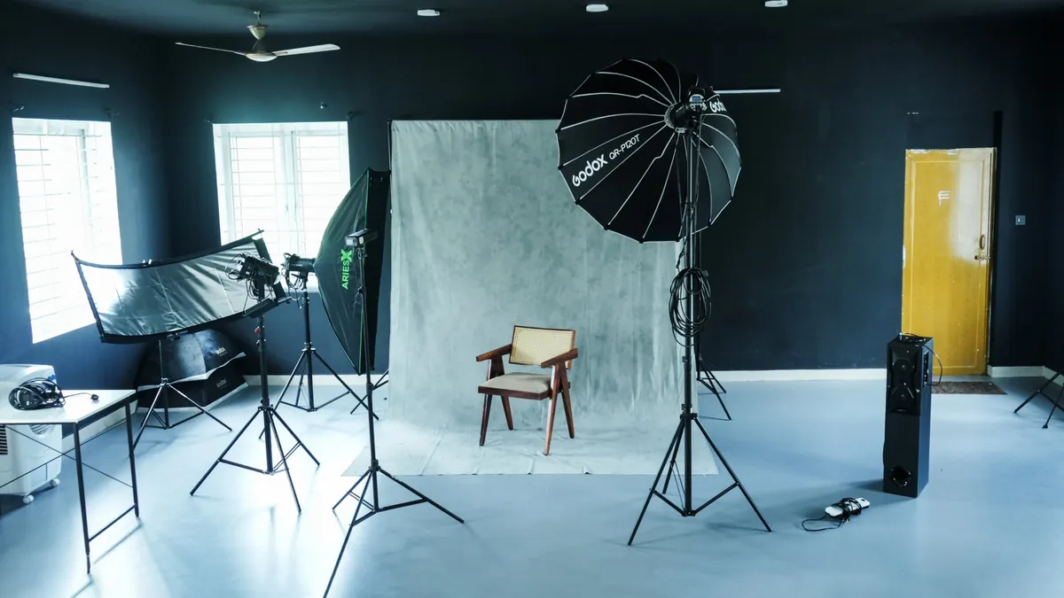 The studio's dark-walled shooting floor: a painted grey backdrop with a cane chair, a large parabolic softbox, a strip softbox, a curved reflector and light stands, with daylight from two windows.